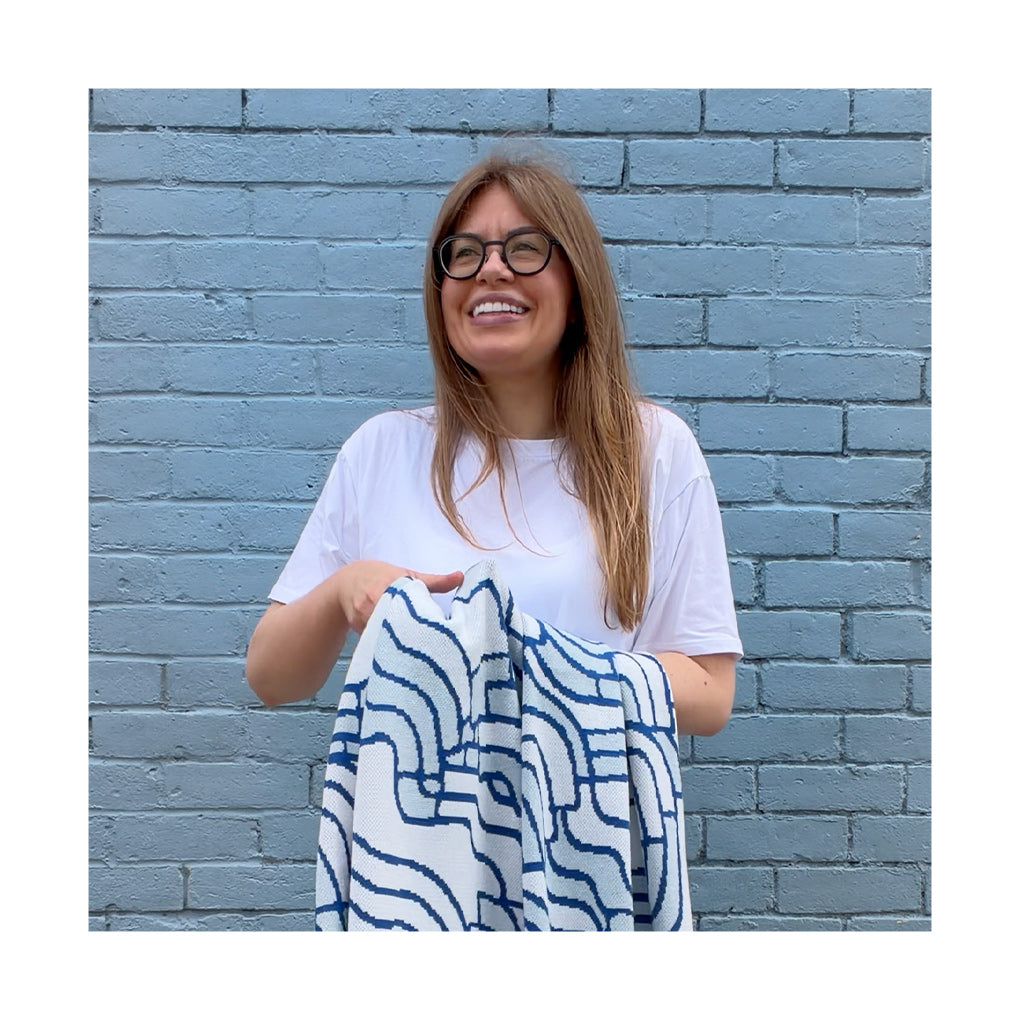 A woman with glasses and a white t-shirt stands in front of a blue brick wall, holding up a white towel or blanket with a blue wavy geometric pattern.